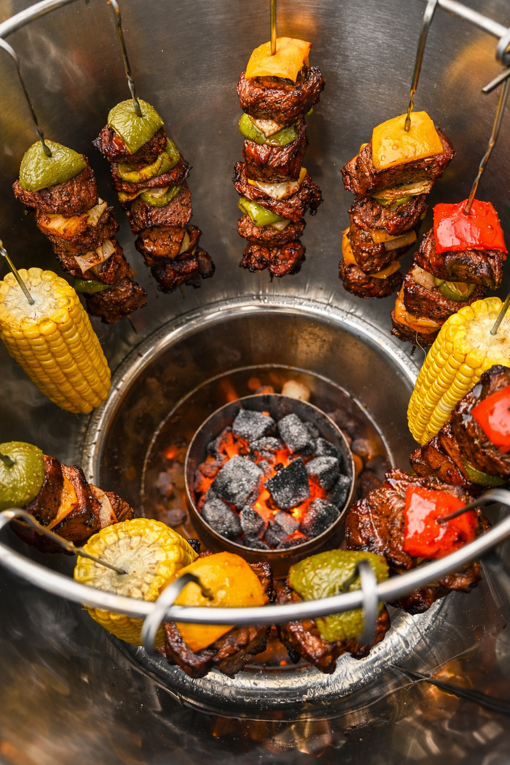 Skewers of meat and vegetables over a small fire pit on a metal surface in stainless steel barrel charcoal smoker